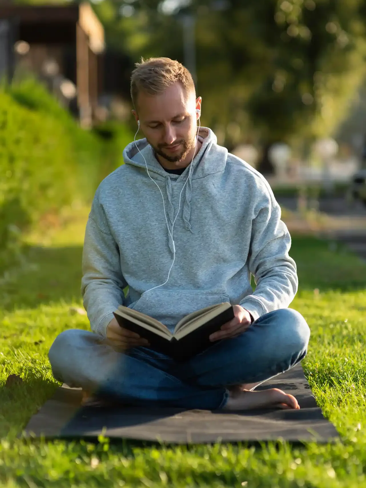 Willkommen auf hausbauplan.com 7 Mann sitzt im Park auf einer Matte und liest ein Buch mit Kopfhörern bei sonnigem Wetter